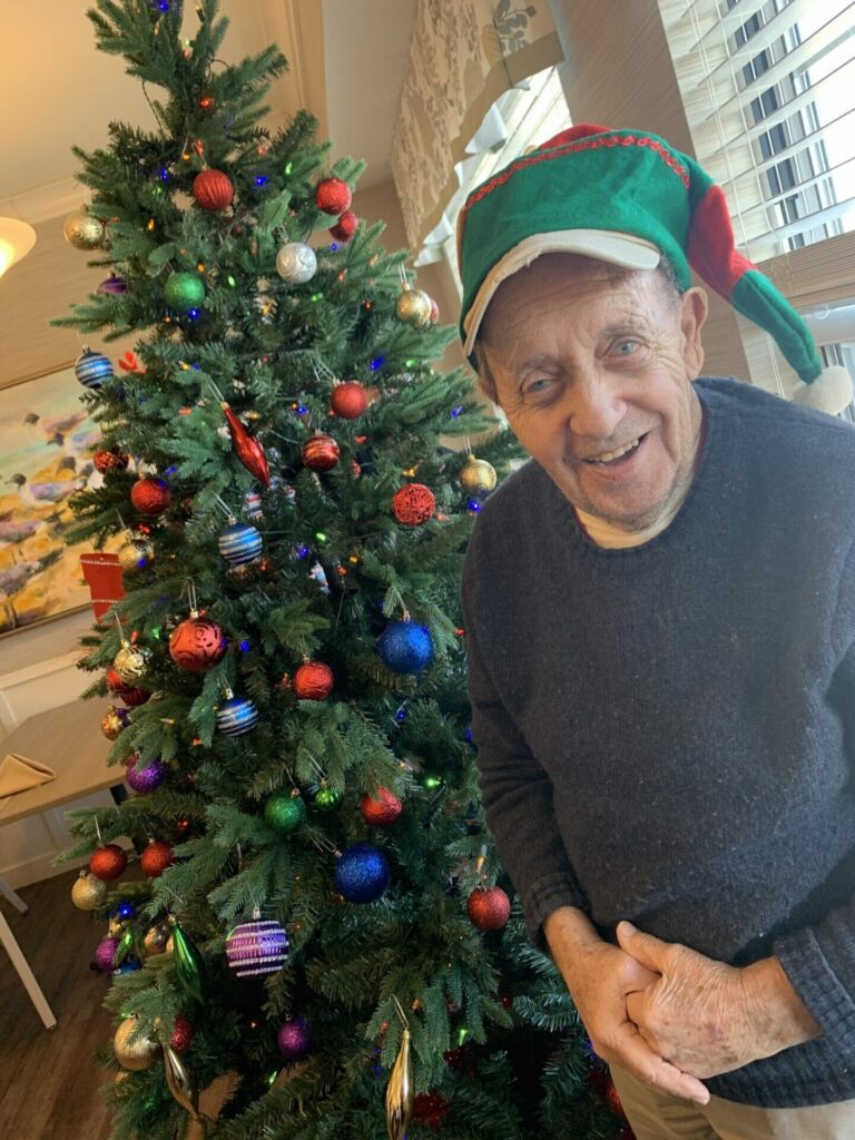 An elder man in blue standing next to a Christmas' tree