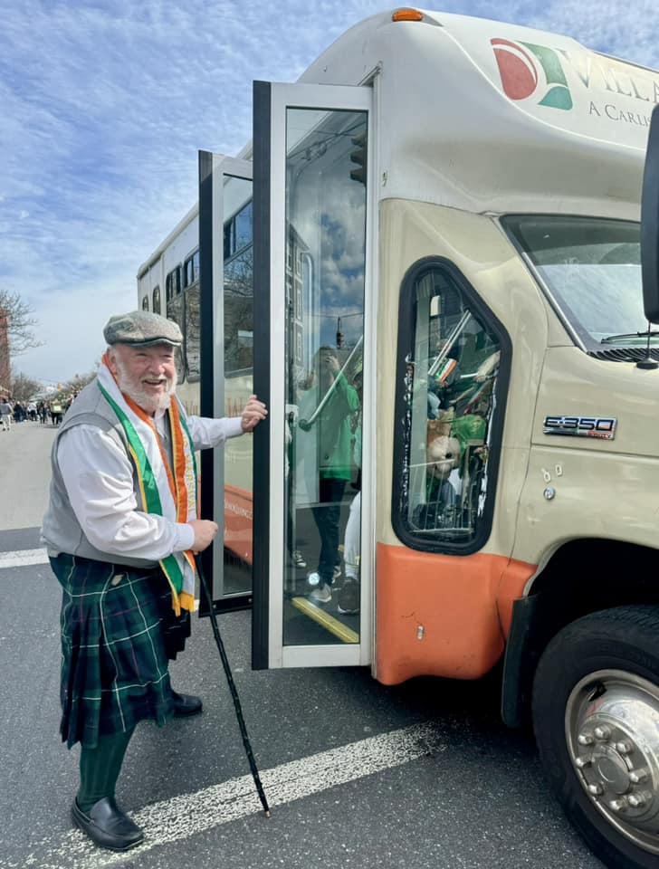 A man in front of a memory  care facility smiling and about to board a bus owned by Village Walk Senior Living