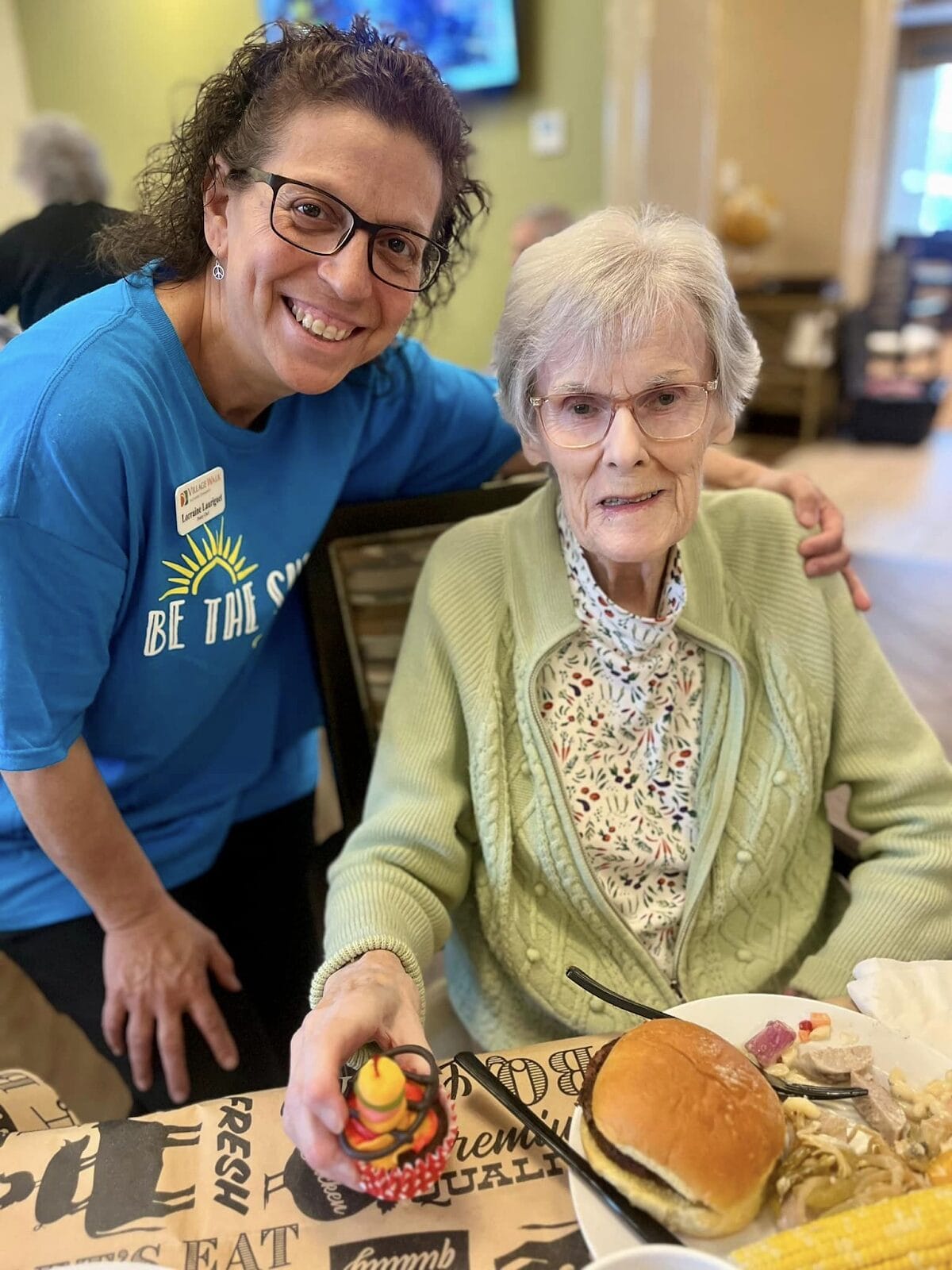 A woman smiling with her arm around an elderly person in a memory care facility