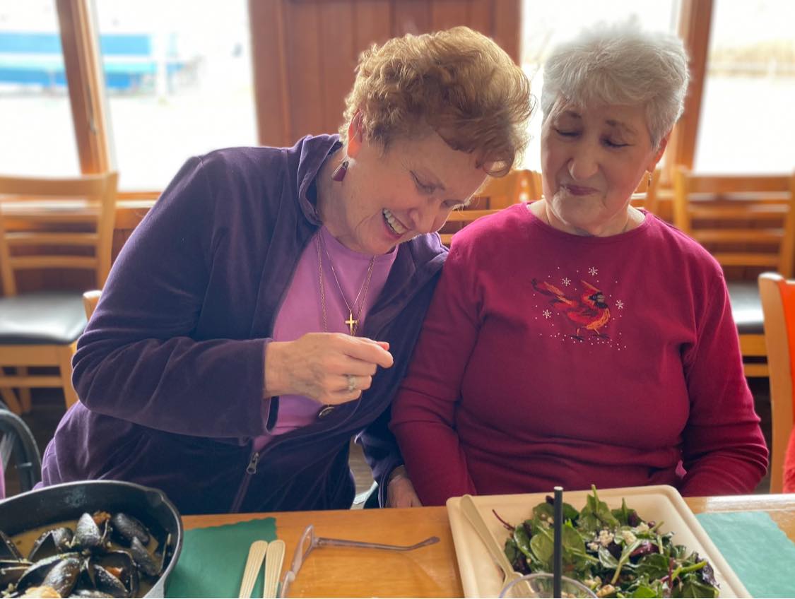 A memory care resident smiling, closing her eyes in laughter, and eating food with a friend of hers at a restaurant