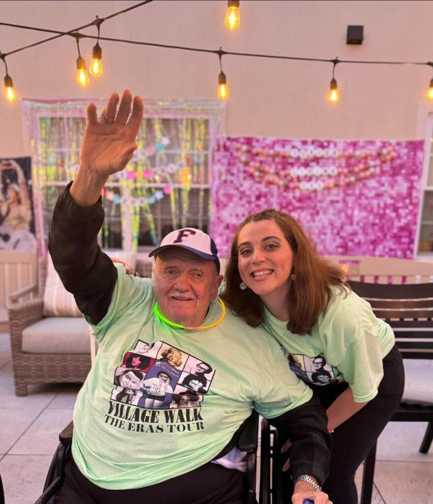 A Village Walk staff member and an elderly man waving at the camera with a lighted scenery backdrop