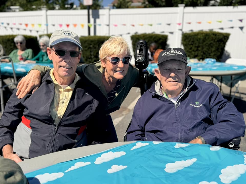 Two elderly men smiling with a woman in the middle between the two men.