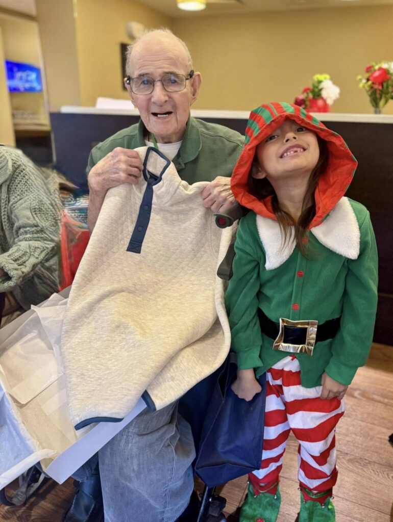 A picture of an elderly man opening a gift of a button down shirt with an elementary school-aged child smiling in Christmas  clothes next to him