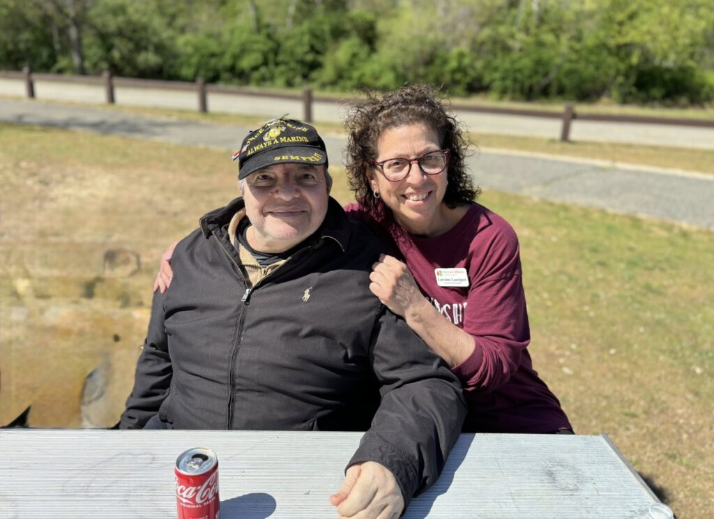 An assisted living facility team member with their arms around a man wearing a veterans hat