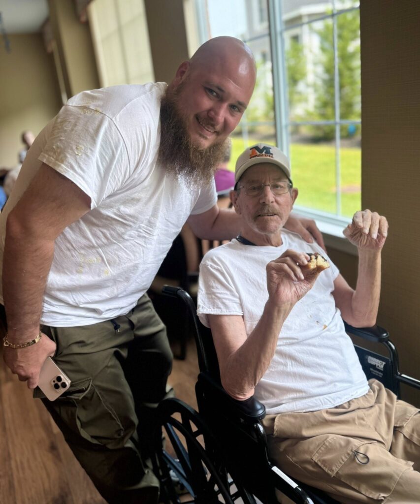 An elderly individual in a wheelchair with a younger adult smiling in an assisted living facility