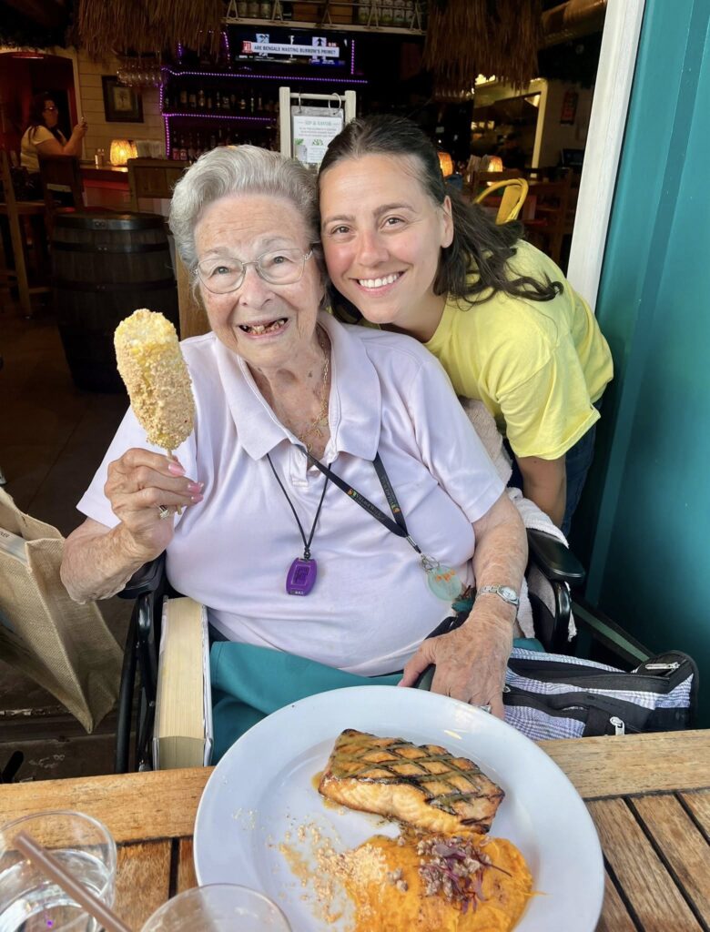 A woman eating a filet of salmon with an assisted living staff member smiling in the background